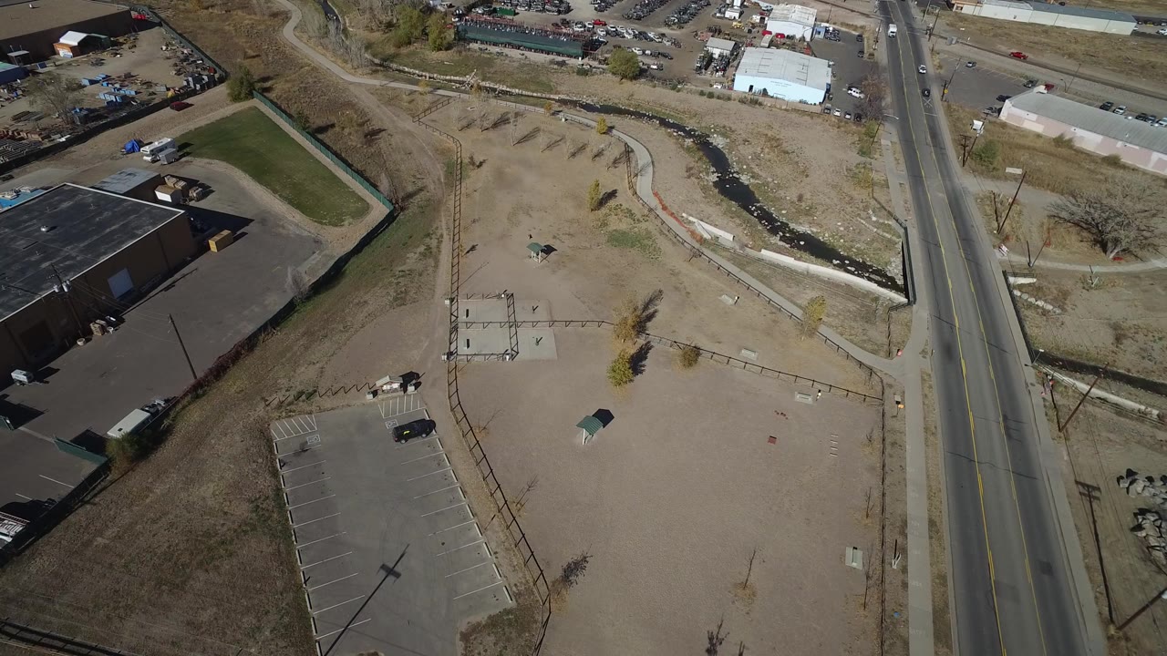 Opening aerial frame over Westminster, Colorado shows a two-lane road beside a parking lot, a low commercial building, dry open ground, and no visible people.