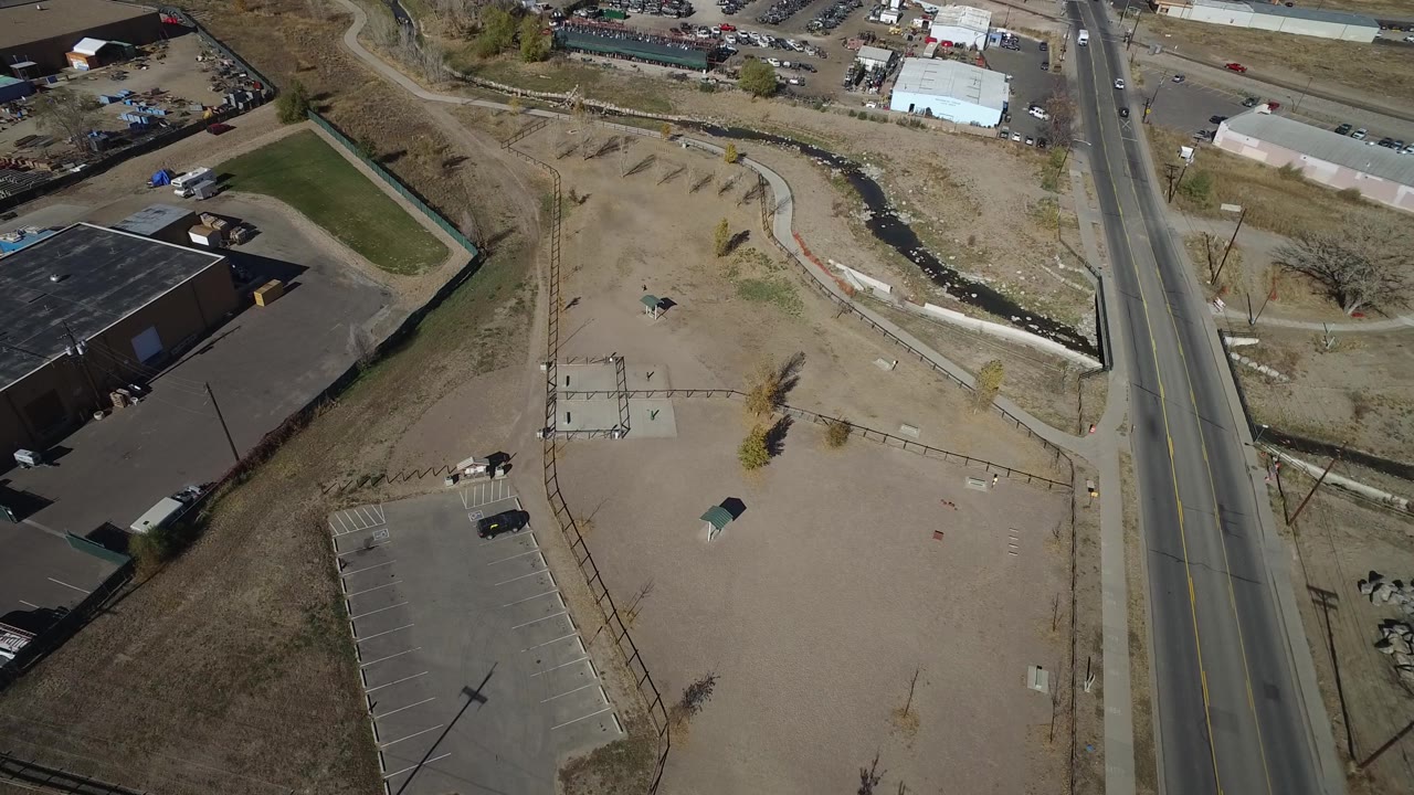 A later aerial frame in Westminster, Colorado keeps the road near the right edge while the fenced parking area, dry ground, and drainage channel fill the center, with no visible people.