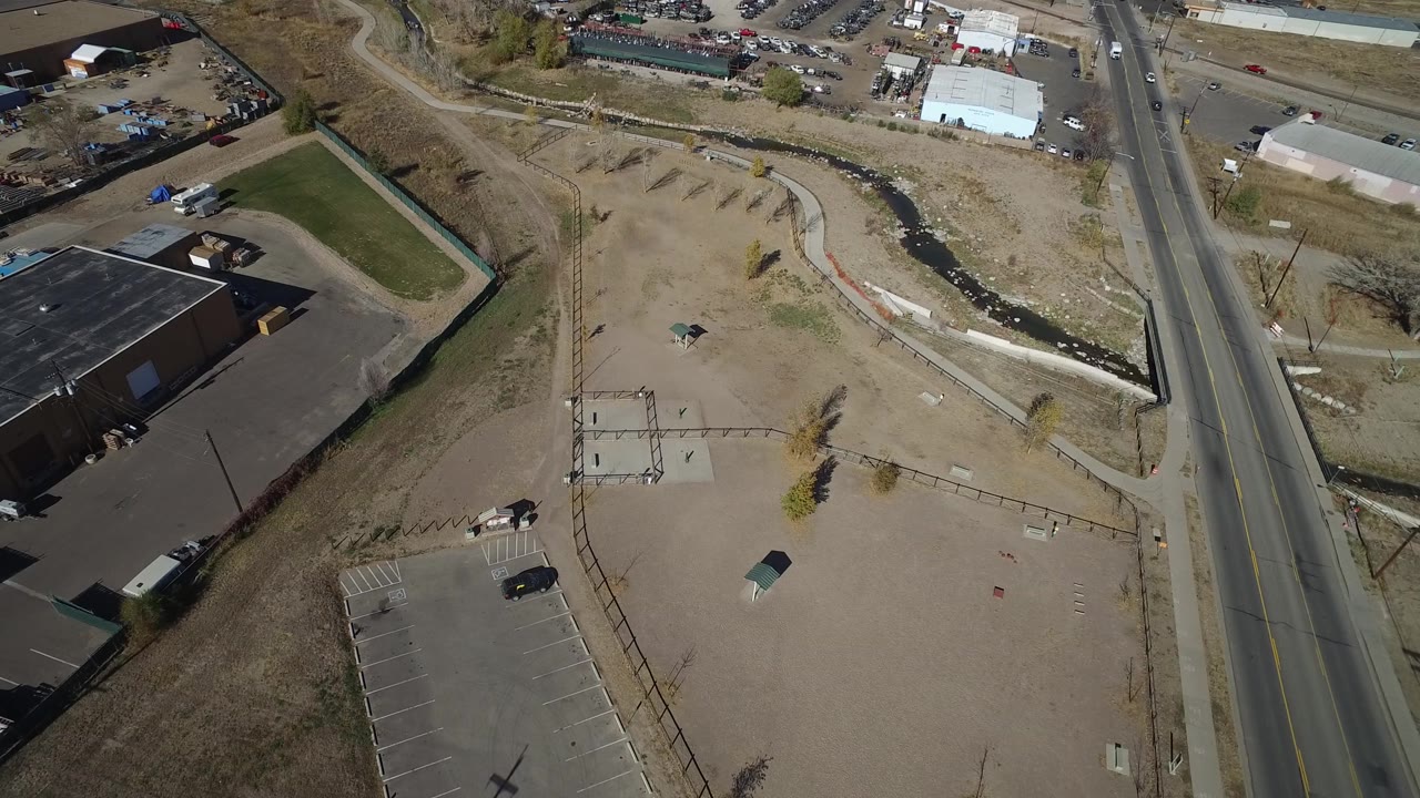 Aerial frame with road, dark-roofed building, parking lot, and open dirt area.