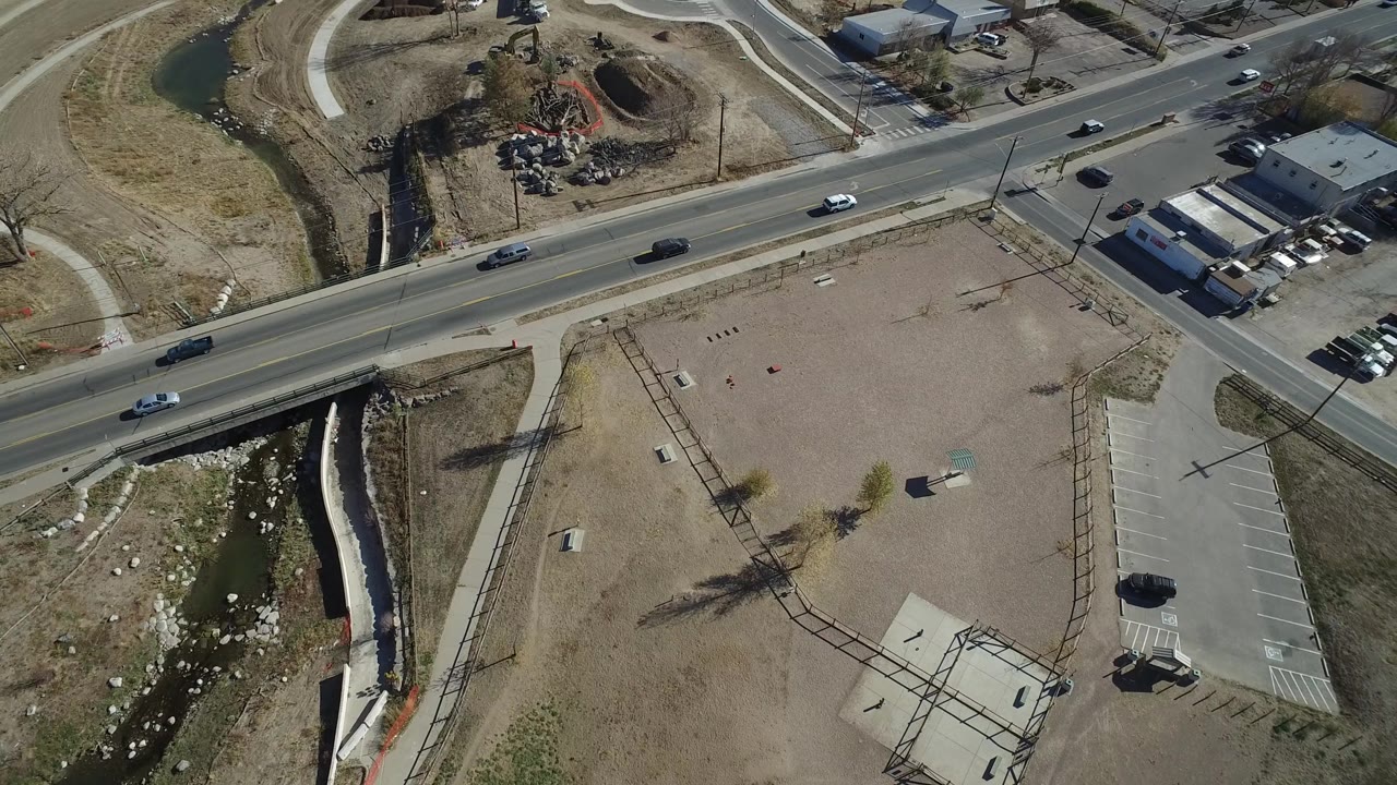 Aerial view in Westminster, Colorado with cars traveling along a paved road, a gravel lot below, a narrow drainage channel at left, commercial buildings at top right, and no visible people.