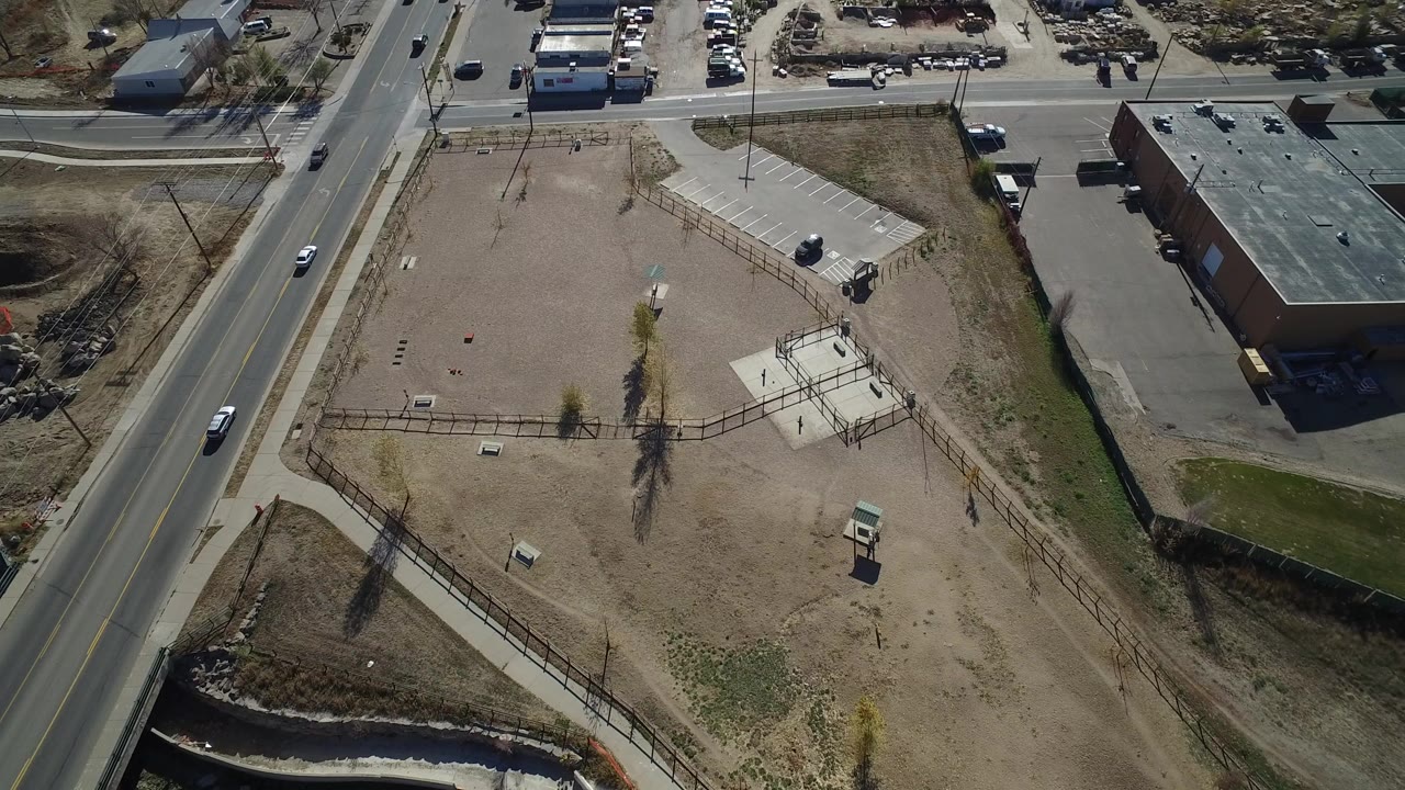Aerial view in Westminster, Colorado, looking down on a fenced dirt field, roadside lanes, a low brick building, parking areas, and no visible people.