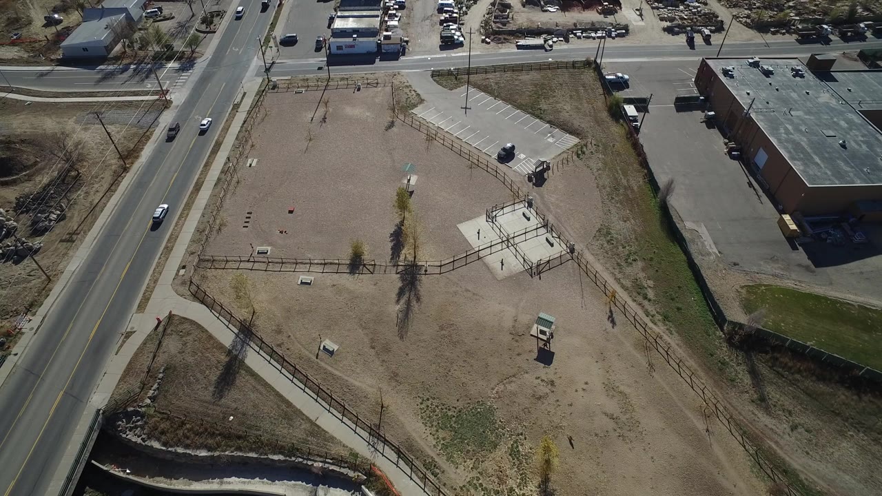 Drone view of a bare fenced lot with a road, sidewalk, and commercial roof nearby.