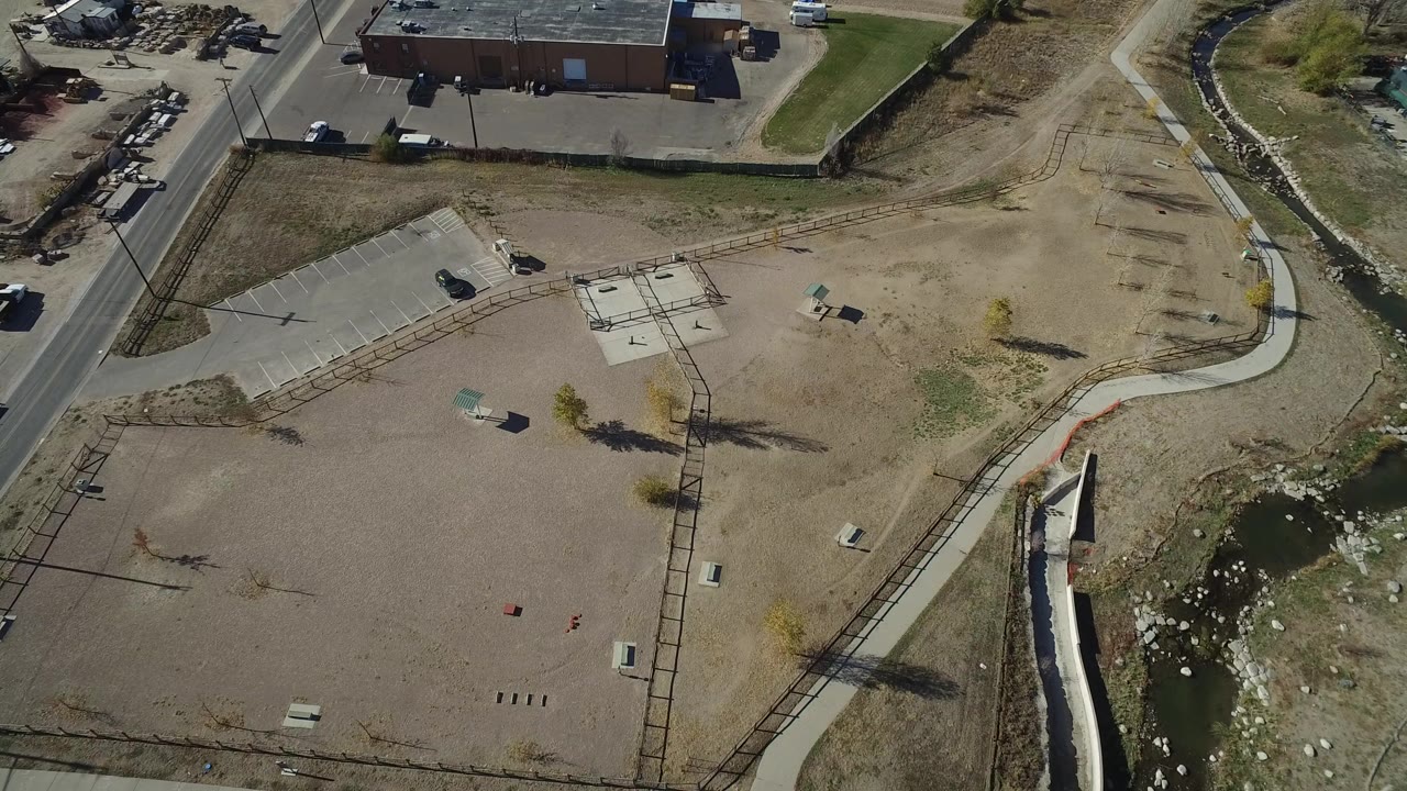Aerial frame over an empty dirt lot in Westminster, Colorado, with a curved sidewalk, narrow drainage channel, road, industrial building, parked cars, and no visible people.