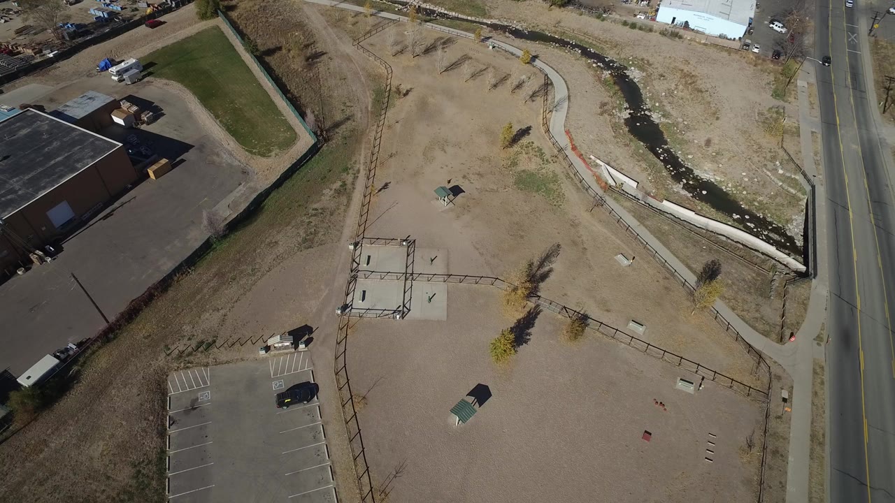 High aerial view in Westminster, Colorado showing a small tan lot, marked parking spaces, a roadside lane, a creek channel, and no visible people.