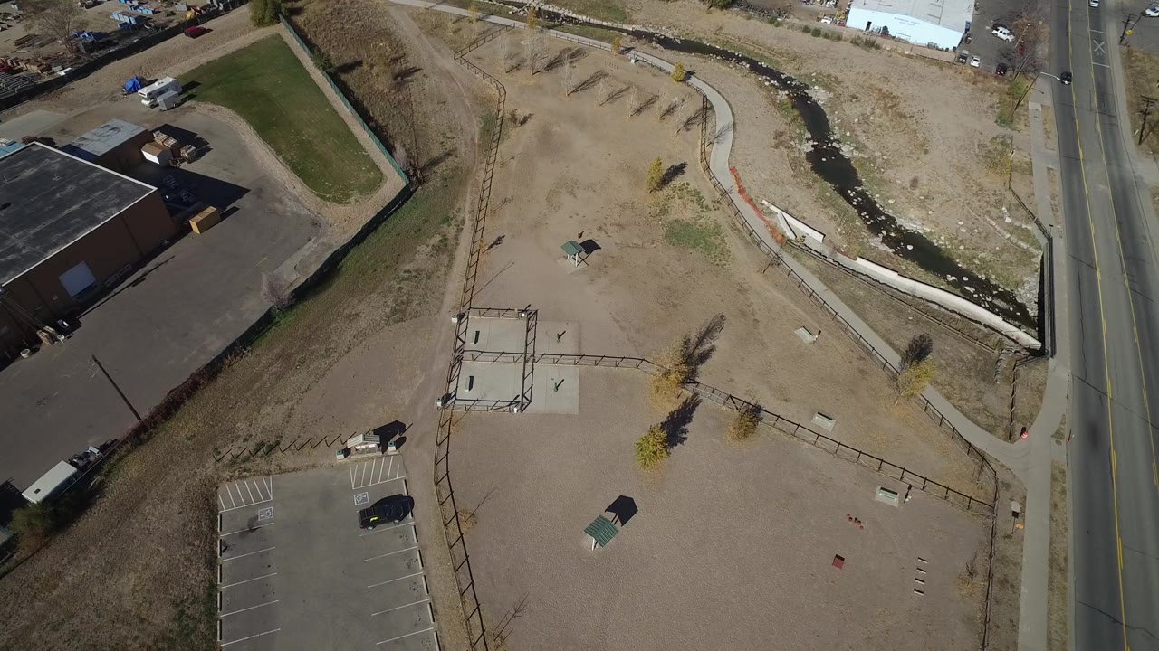 Aerial frame in Westminster, Colorado with the empty dirt lot centered, asphalt parking bays at lower left, the road on the right edge, and no visible people.