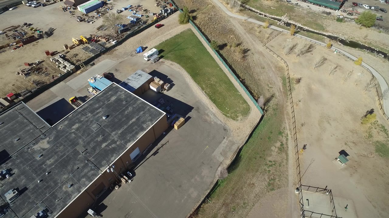 Aerial frame over Westminster, Colorado with the black roof filling the lower left, gravel storage areas and parked vehicles above, and a narrow maintained grass strip at right with no visible people.