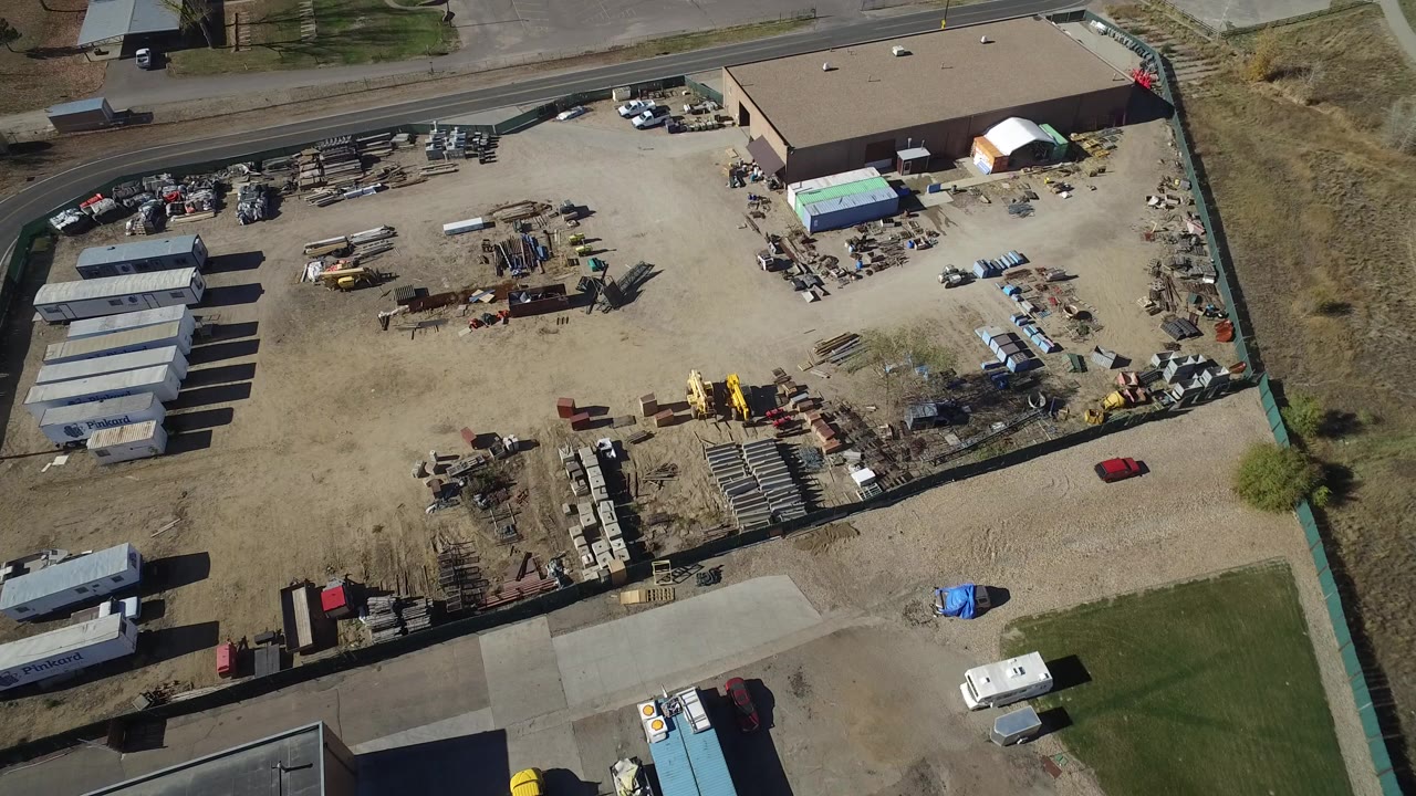 Aerial frame over a Westminster, Colorado industrial yard with storage sheds on the left, a large warehouse at the upper right, parked vehicles, scattered materials, and no visible people.