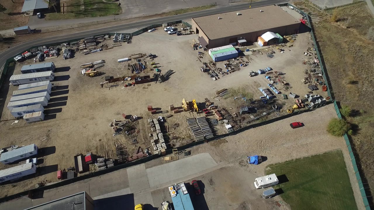 A high view of the Westminster industrial lot shows the road along the top edge, rows of sheds on the left, equipment and material piles in the center, and no visible people.