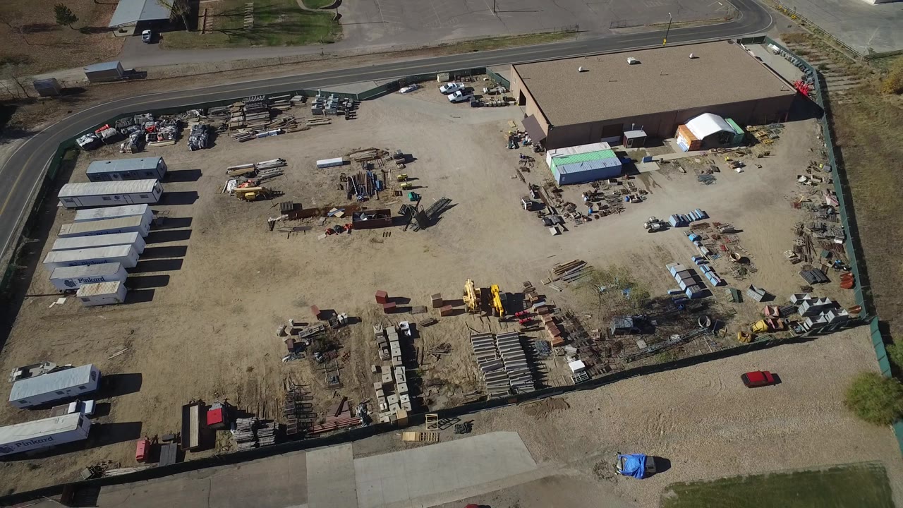 Aerial industrial yard with a large warehouse, parked trucks, sheds, and stacked supplies.