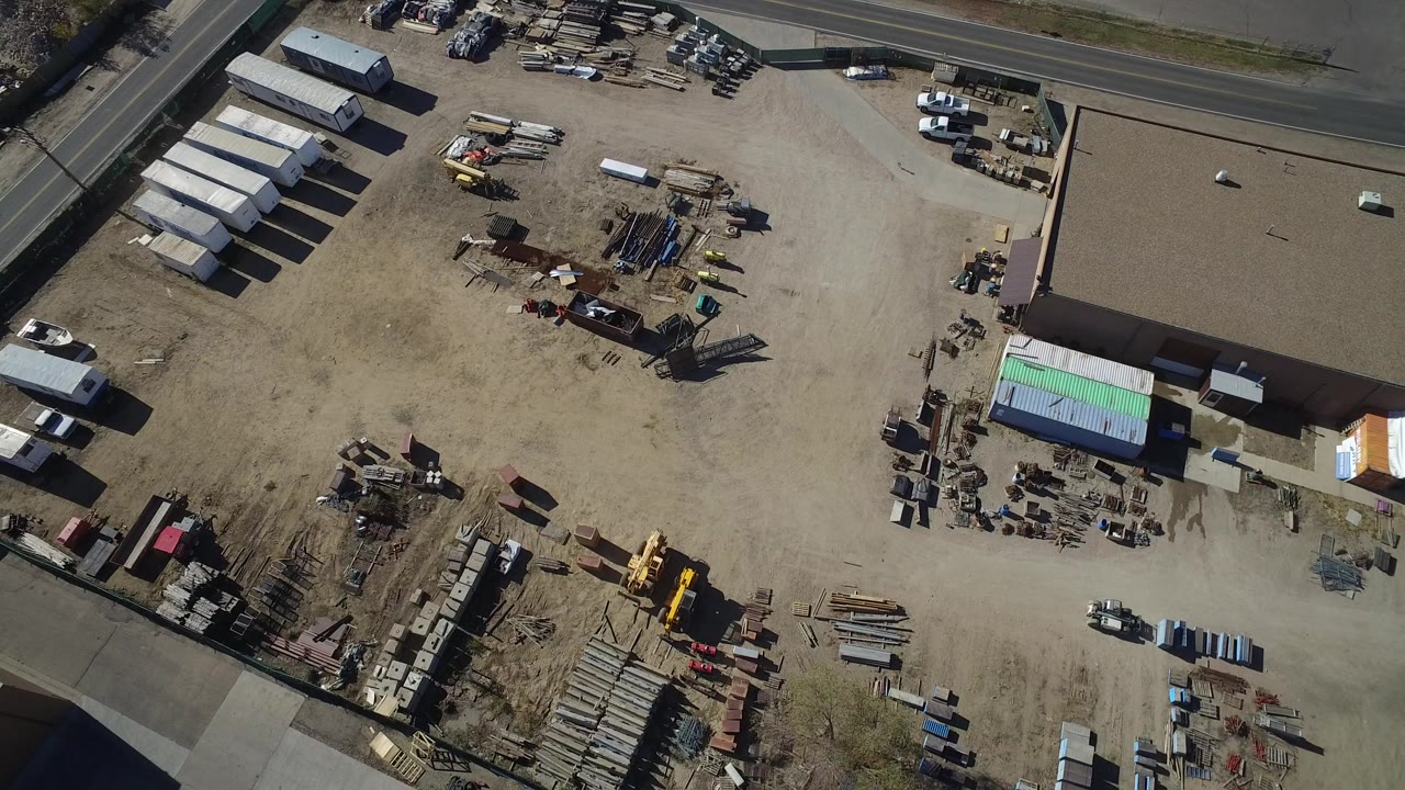 Aerial frame over a Westminster, Colorado industrial yard with shipping containers, scattered materials, warehouse roofs, a road at the top edge, and no visible people.