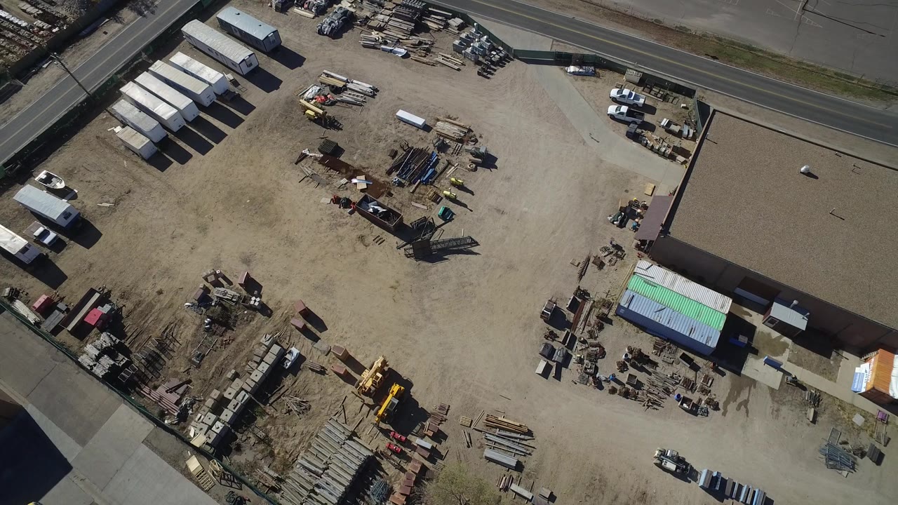 Overhead industrial lot with stacked materials, yellow machinery, containers, and adjoining road.