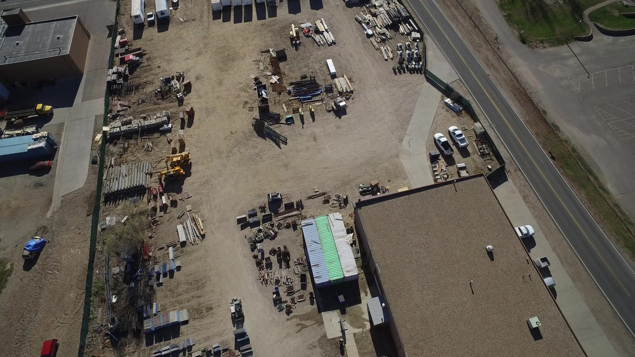 Overhead view of a dusty yard with sheds, parked vehicles, stacked materials, and a road.