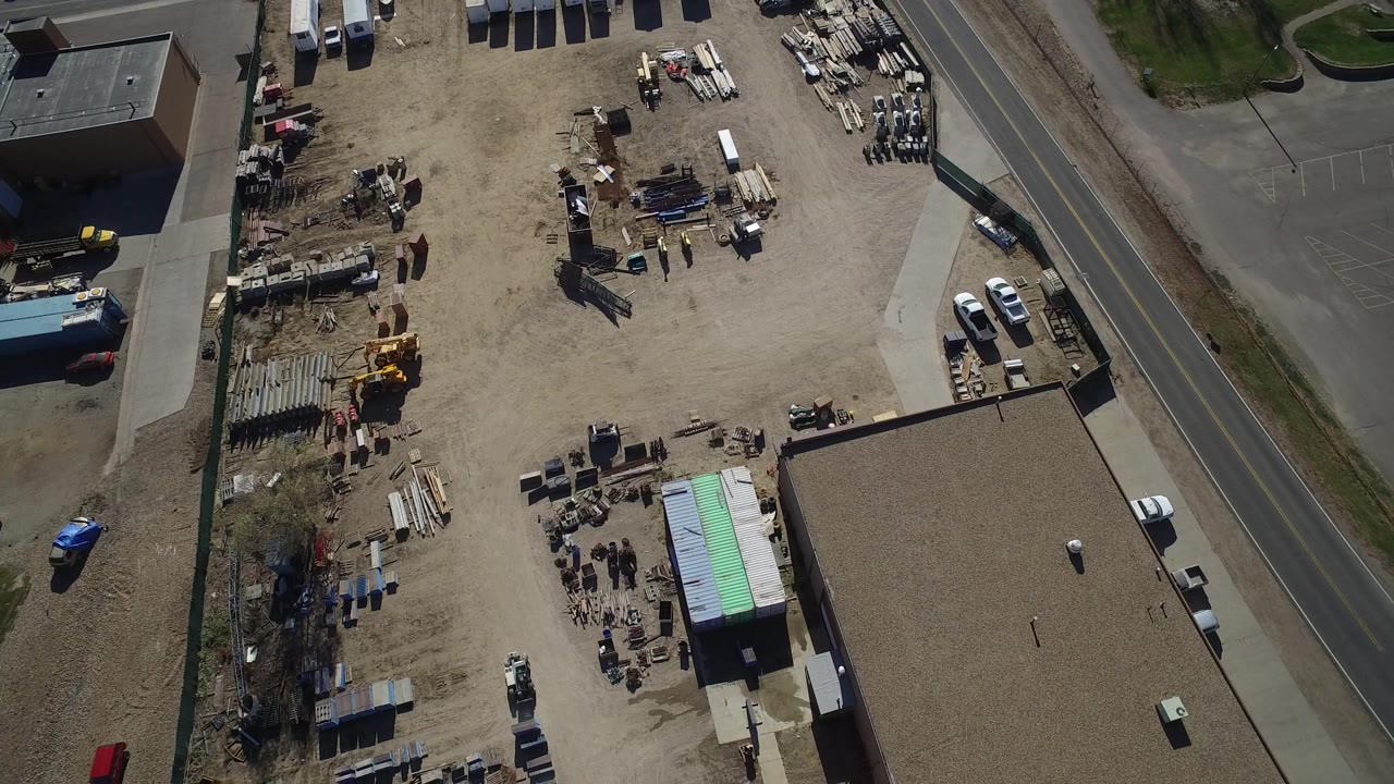 A second aerial frame in Westminster, Colorado keeps the yard centered, with piles of materials, a green-roofed shed, parked vehicles near the road, and no visible people.