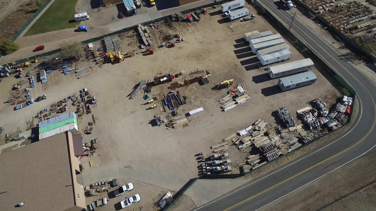 Overhead drone-style view of a fenced construction yard in Westminster, Colorado, with stacked materials, trailers, parked vehicles, a tan-roofed building edge, and no visible people.