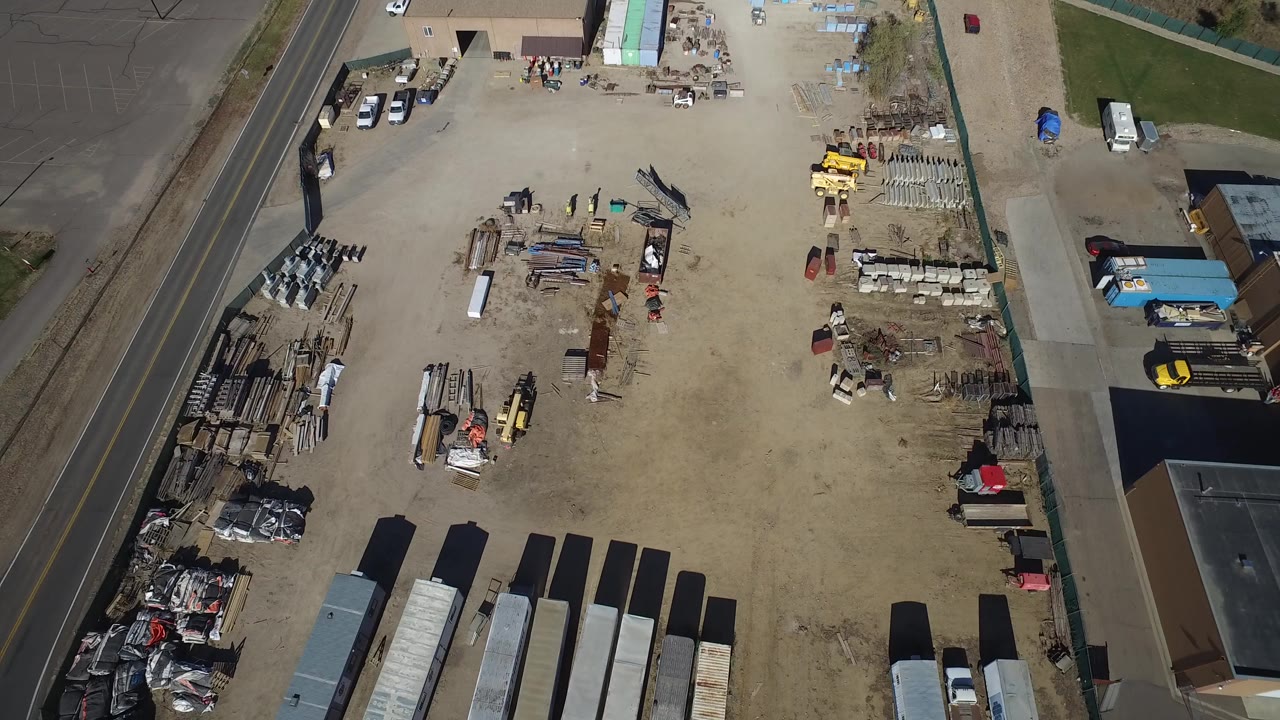 Overhead daylight frame of a sandy industrial yard in Westminster, Colorado, with stacked materials, trailers, a road at left, low buildings at the edges, and no visible people.