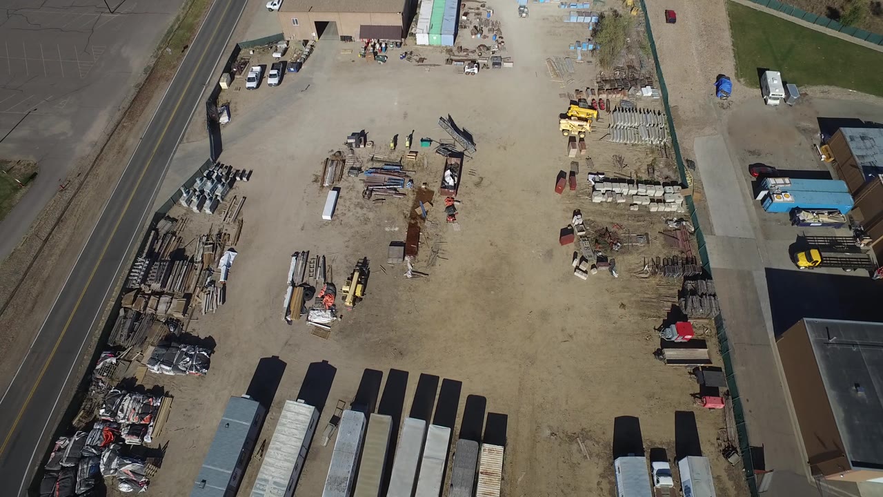 Aerial frame of the Westminster storage yard showing long trailers at the lower edge, scattered pipes and pallets near the center, a road along the left side, and no visible people.