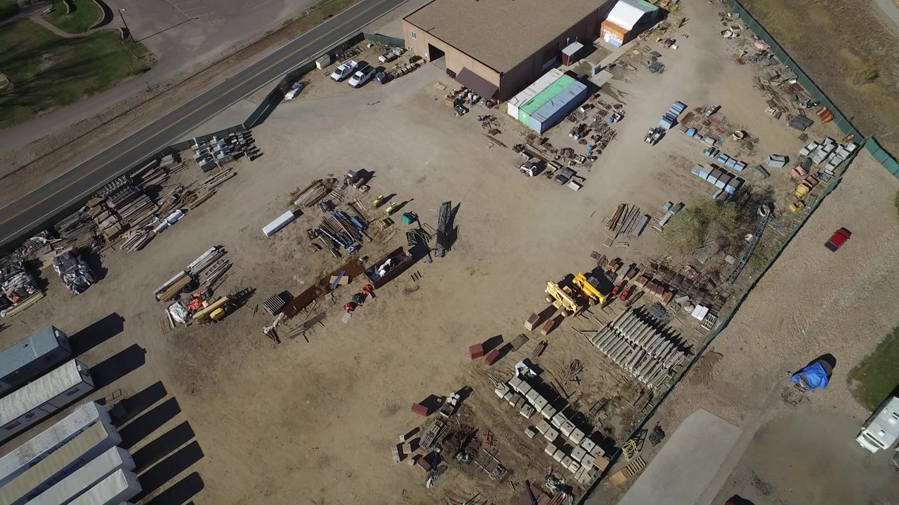 Aerial frame over the Westminster business lot showing a tan dirt surface, semi trailers near the lower right, yellow machinery near the center, stacked materials, parked vehicles, and no visible people.