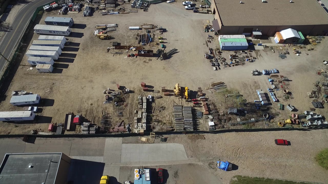 High drone view of the Westminster construction yard with rows of trailers on the left, piles of materials in the center, a red car near the lower road, and no visible people.