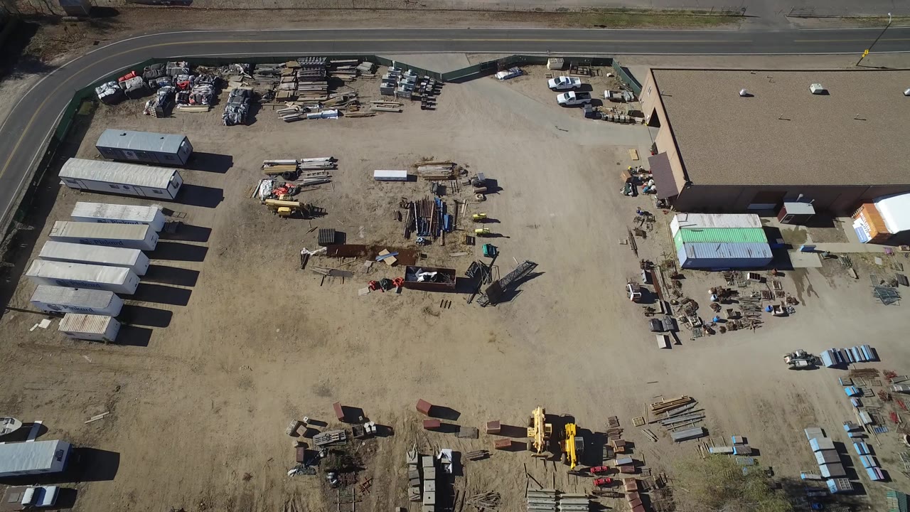 Aerial frame in Westminster, Colorado showing rows of small sheds, scattered equipment, stacked lumber or pipe, a tan warehouse building, parked vehicles, and no visible people.