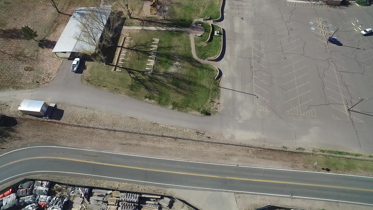 Overhead frame in Westminster, Colorado showing a curved two-lane road, paved parking areas, a white-roofed structure, lawns, tree shadows, and no visible people.