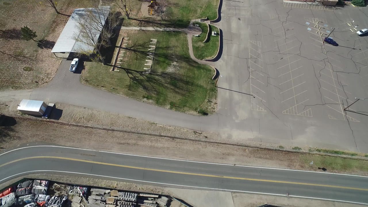 Overhead Westminster, Colorado view with the road along the bottom, empty striped parking spaces to the right, a white-roofed building near center, lawns, trees, and no visible people.