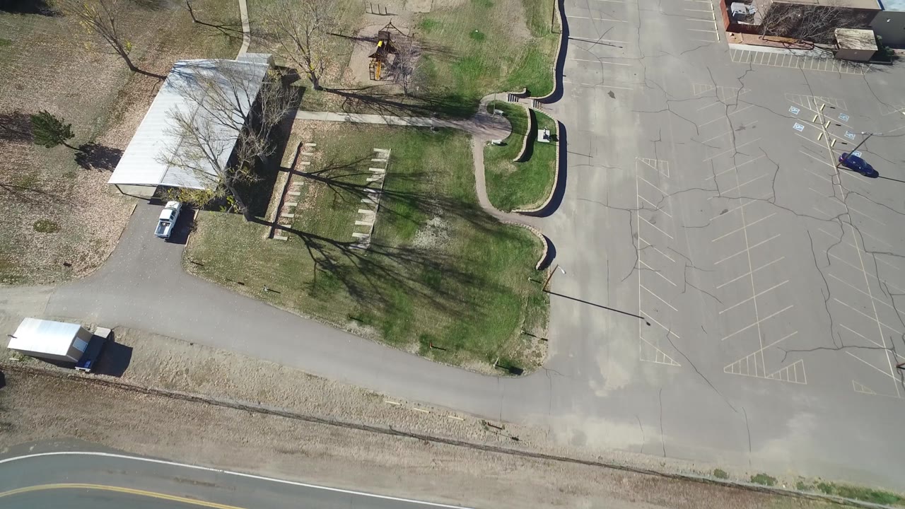 Top-down suburban scene with road, parking lot, lawn, trees, and small structures.