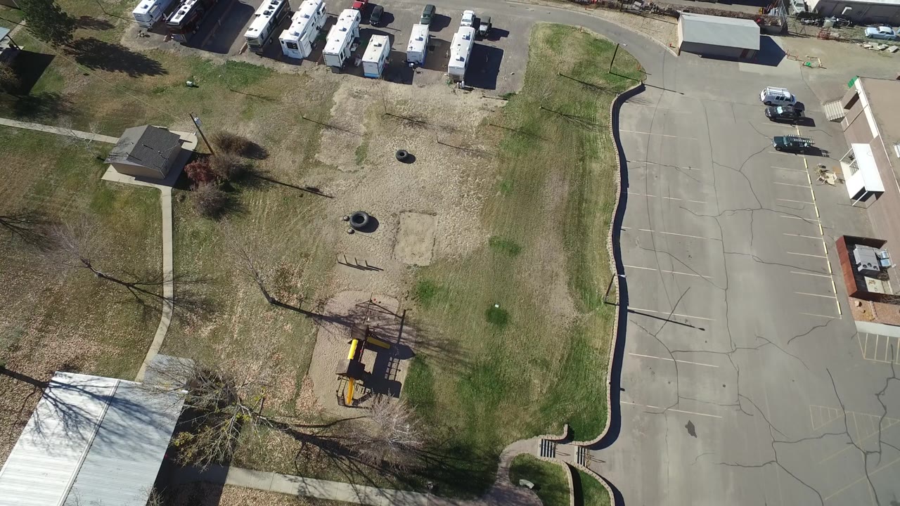 Aerial frame over Westminster, Colorado with a light-roofed warehouse, a row of RVs beside the road, patchy grass, pavement, and no visible people.