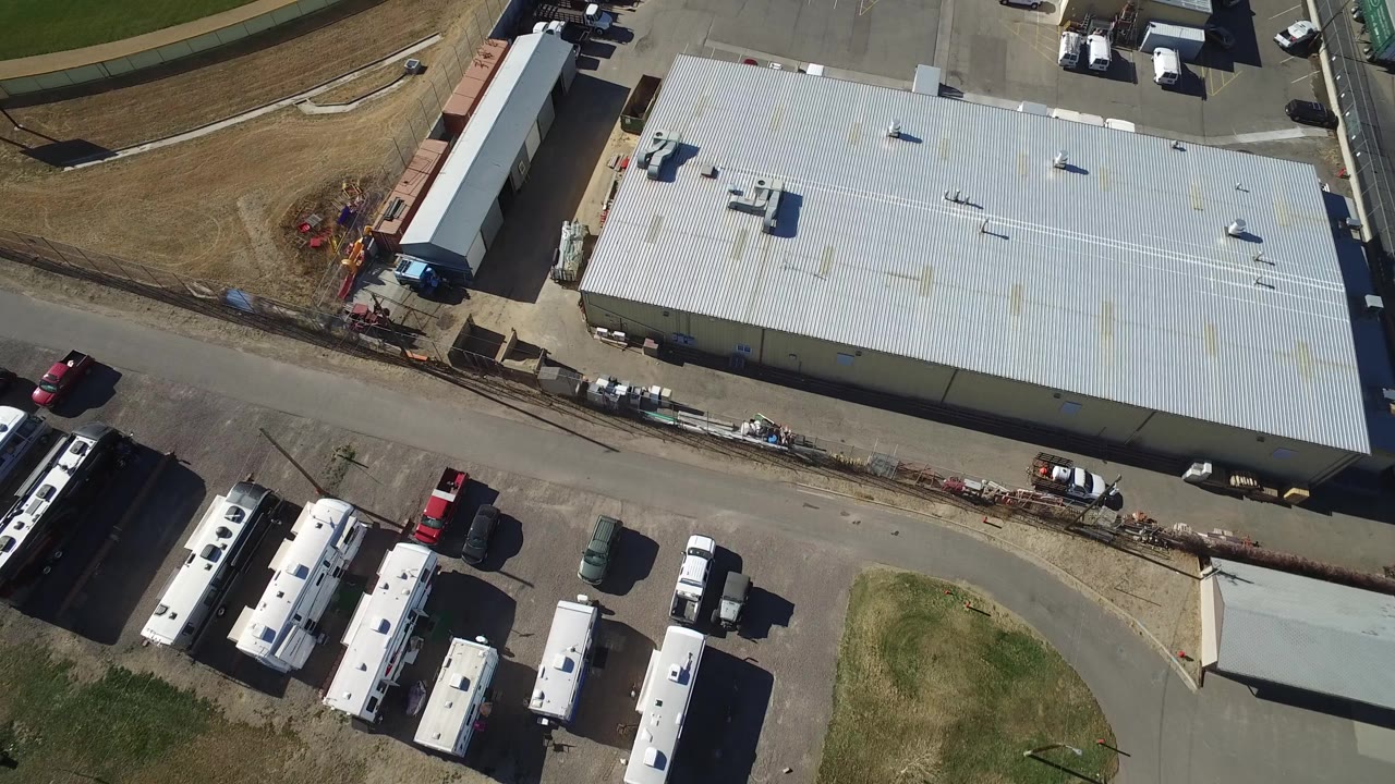 A slightly shifted overhead frame in Westminster, Colorado centers the pale industrial roof with buses, trucks, cars, pavement, grass margins, and no visible people.