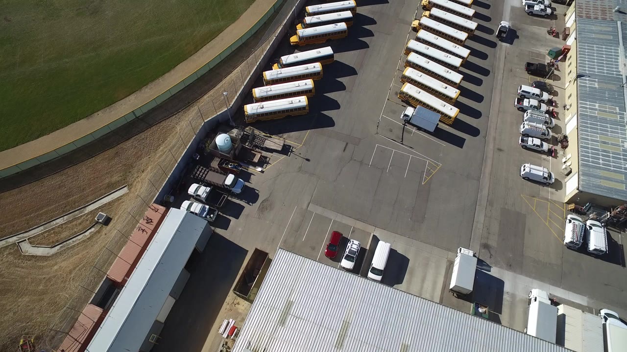 Aerial view in Westminster, Colorado, showing rows of buses, parked cars, a paved terminal lot, and a curved green field edge with no visible people.