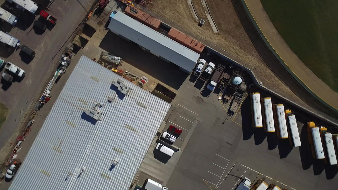 Top-down aerial view of a metal-roofed building, parking lot, buses, and grass.
