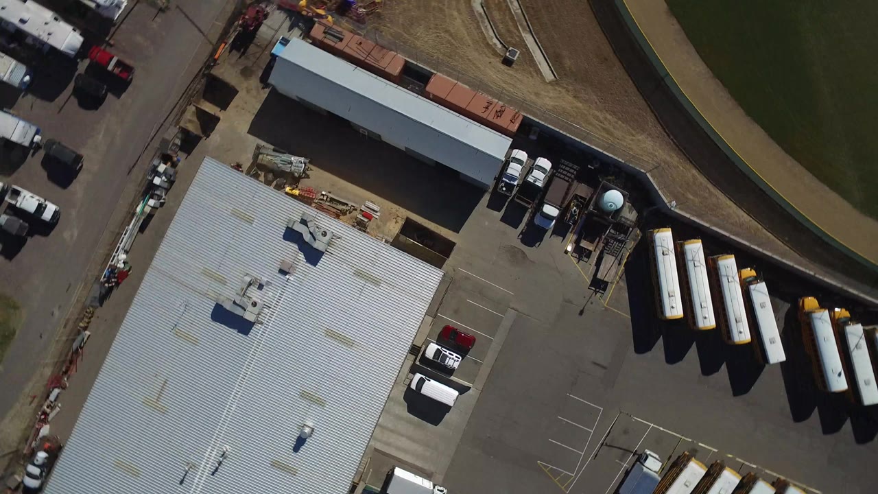 Overhead Westminster, Colorado view with the corrugated roof lower left, parked white trucks near the building, buses or trailers to the right, grass above, and no visible people.