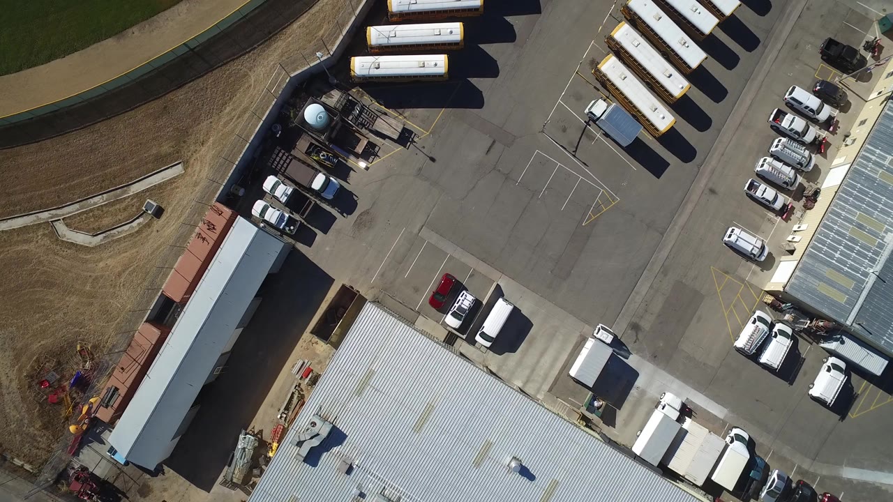 Overhead frame from Westminster, Colorado showing a paved transportation yard with metal-roofed sheds, parked cars, work trucks, rows of white buses or trailers, brown grass, and no visible people.