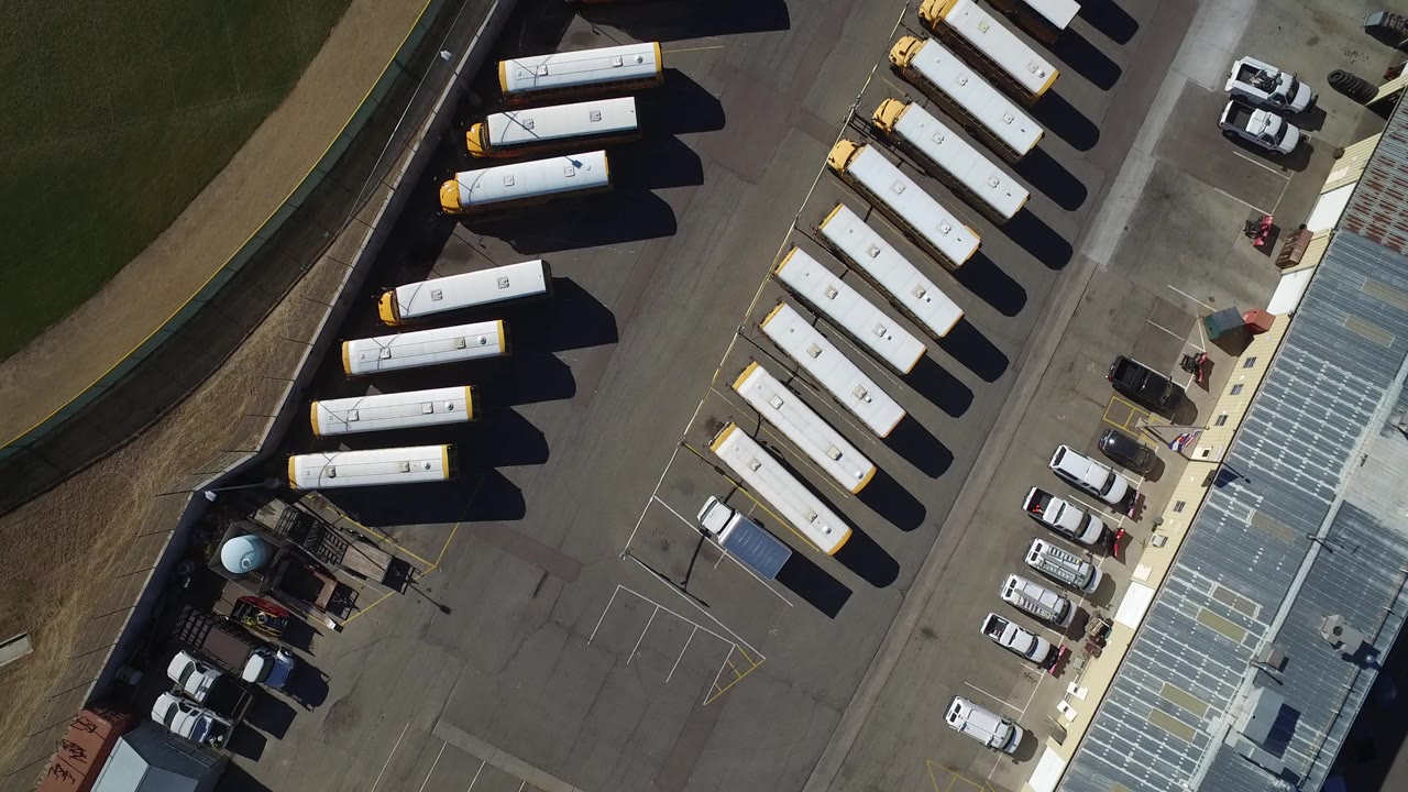 Top-down aerial frame in Westminster, Colorado showing yellow school buses lined along pavement with grass and a curved road at the left, no visible people.