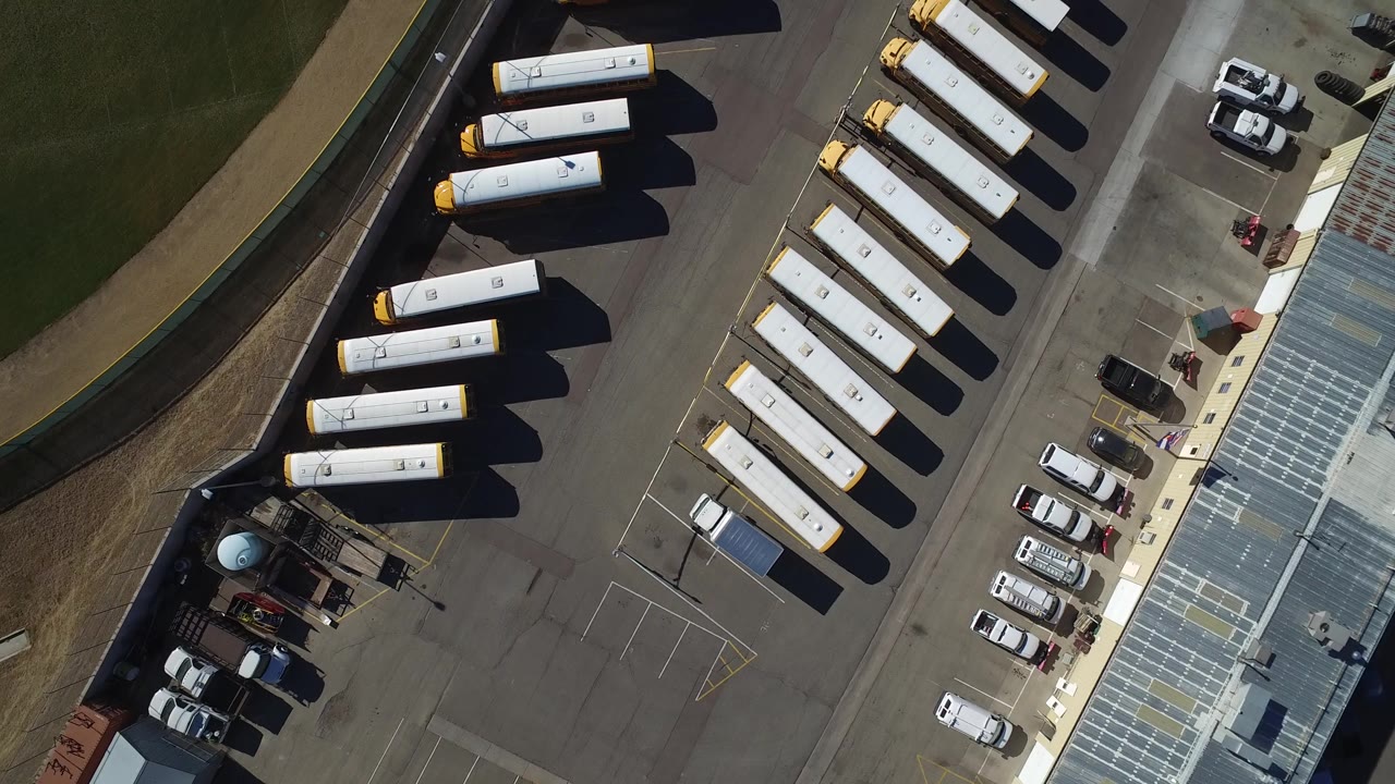 Aerial frame over Westminster, Colorado with two rows of yellow school buses, parked cars near a building edge, and long shadows across the lot, no visible people.