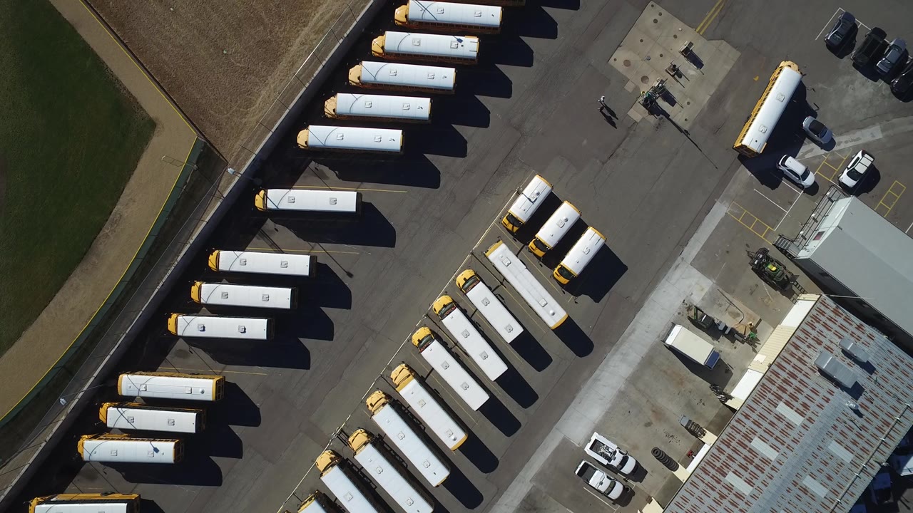 Overhead Westminster, Colorado view with parked goods transport vehicles lined along the asphalt, a low industrial roof at lower right, grass at the edge, and no clearly visible people.