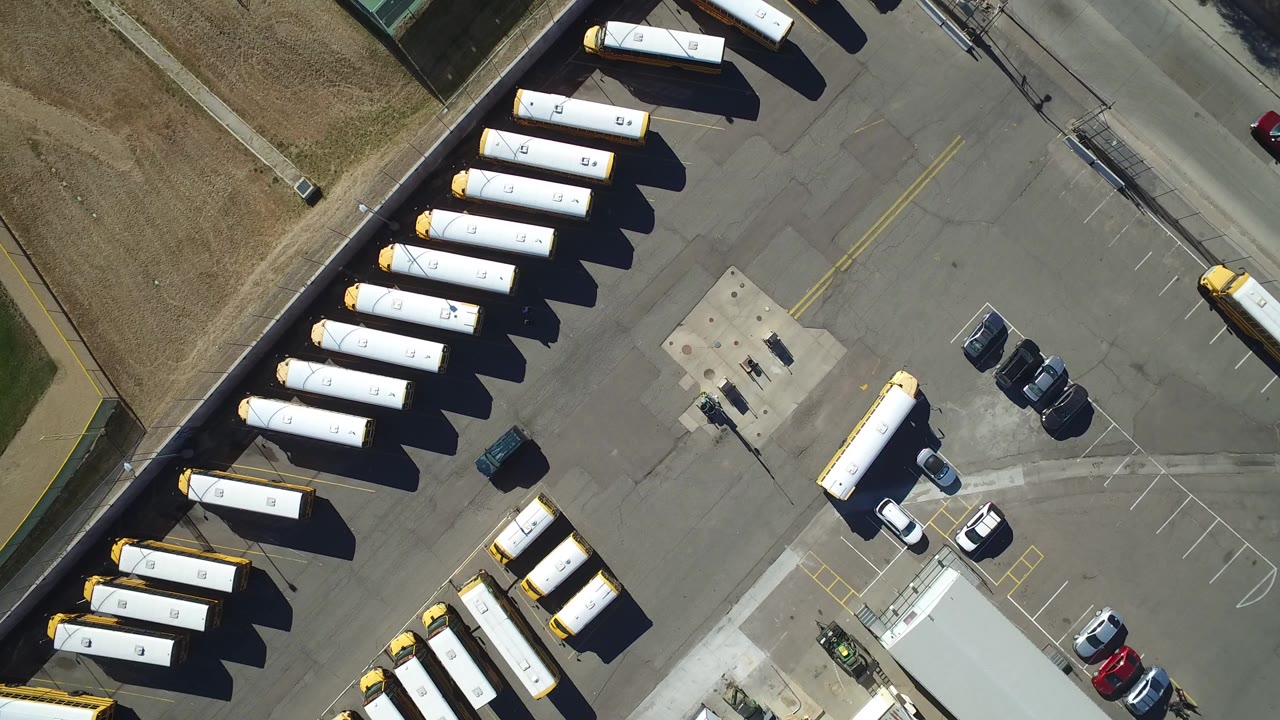 Straight-down aerial frame in Westminster, Colorado showing rows of yellow-and-white buses, a paved parking lot, a road edge, part of a green tennis court, and no visible people.
