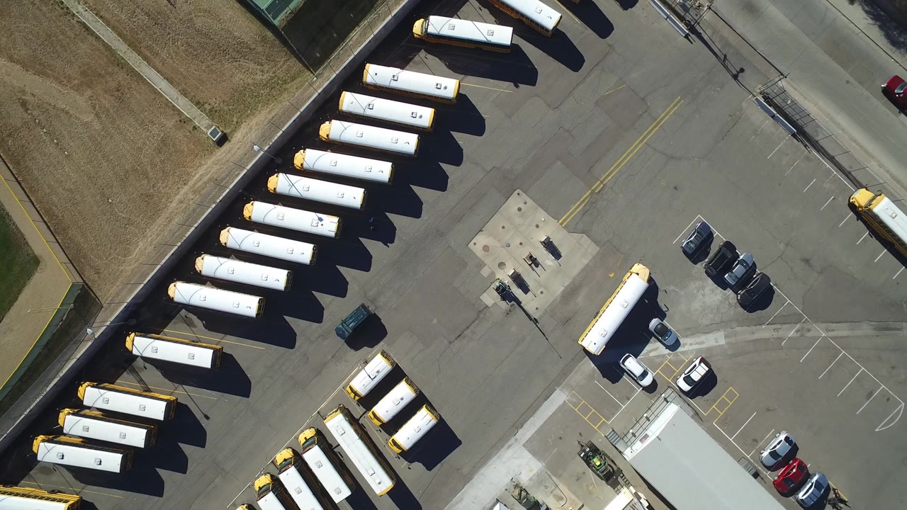 Overhead Westminster, Colorado view with the bus rows shifted slightly across frame, parked cars near the lot entrance, a two-lane road, and no visible people.
