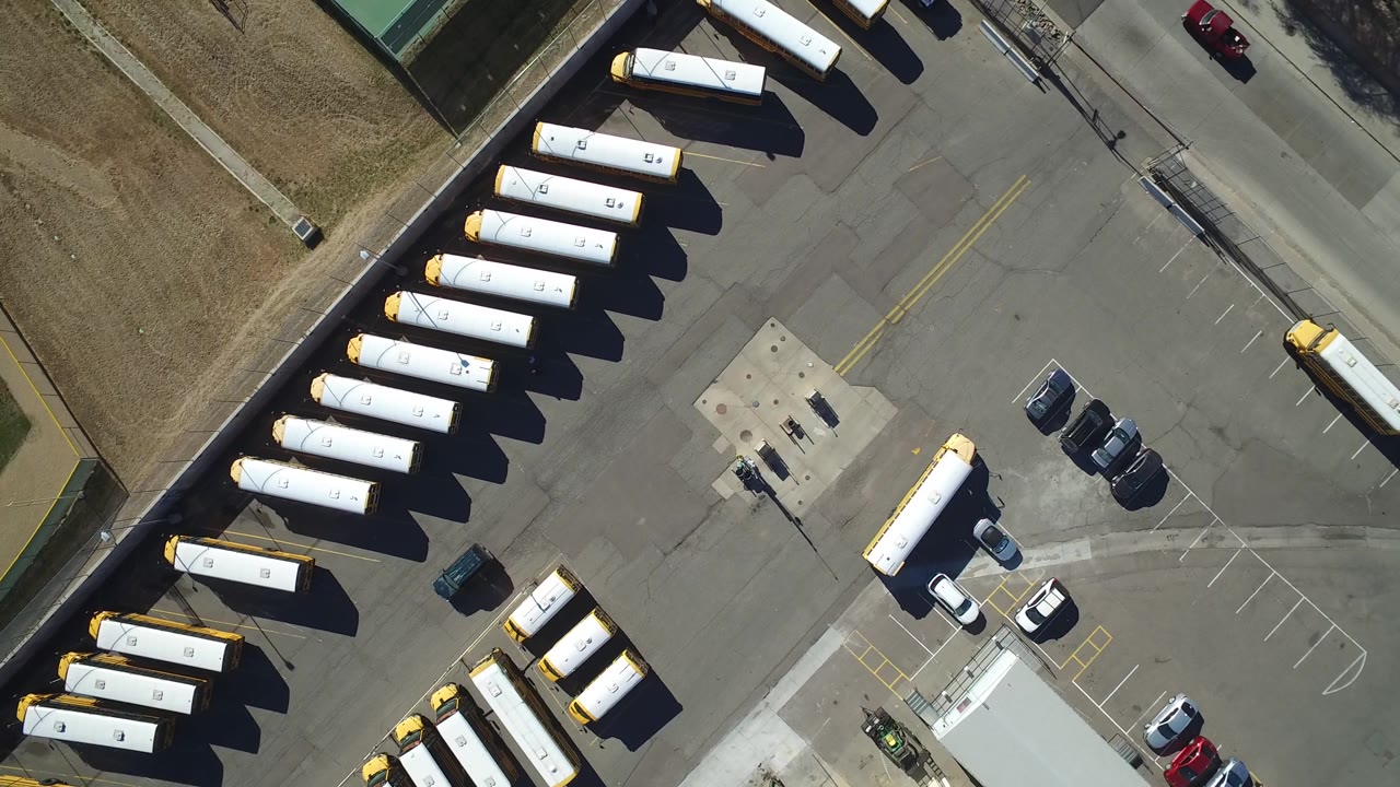 Parking lot overhead with buses, cars, grass, and a road crossing the frame.