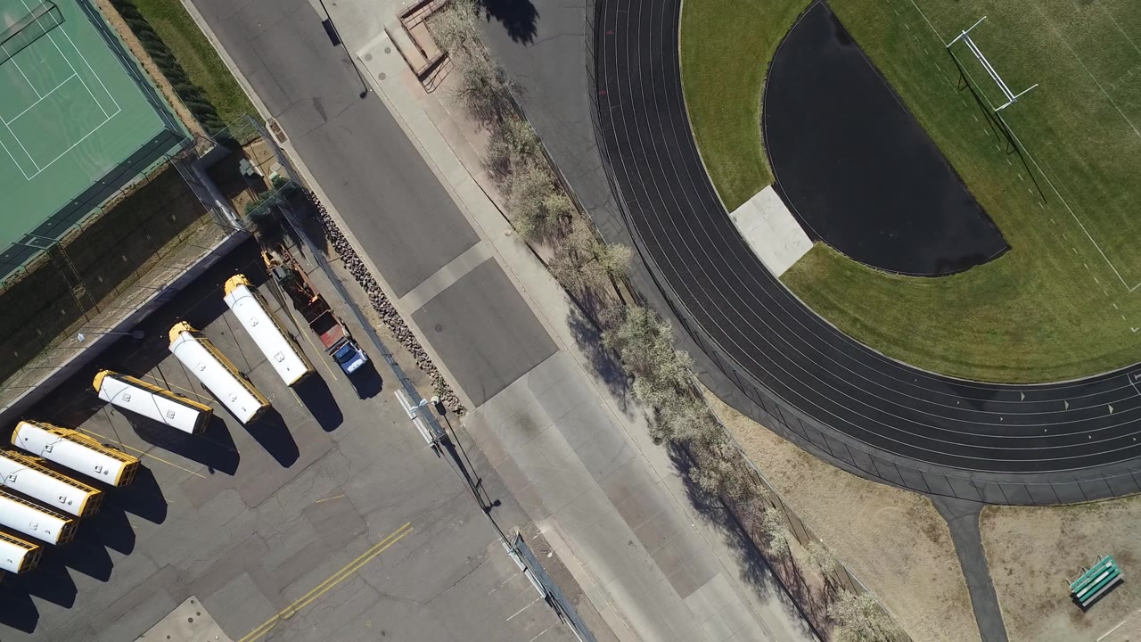 Overhead aerial frame in Westminster, Colorado showing the edge of a green football field, black running track, tennis court, paved road, parked yellow school buses, and no visible people.