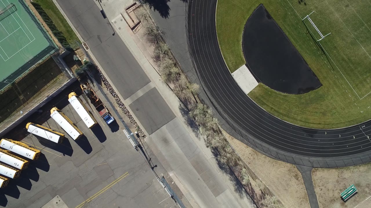 Top-down school athletics area with track, turf field, tennis court, road, and parked buses.