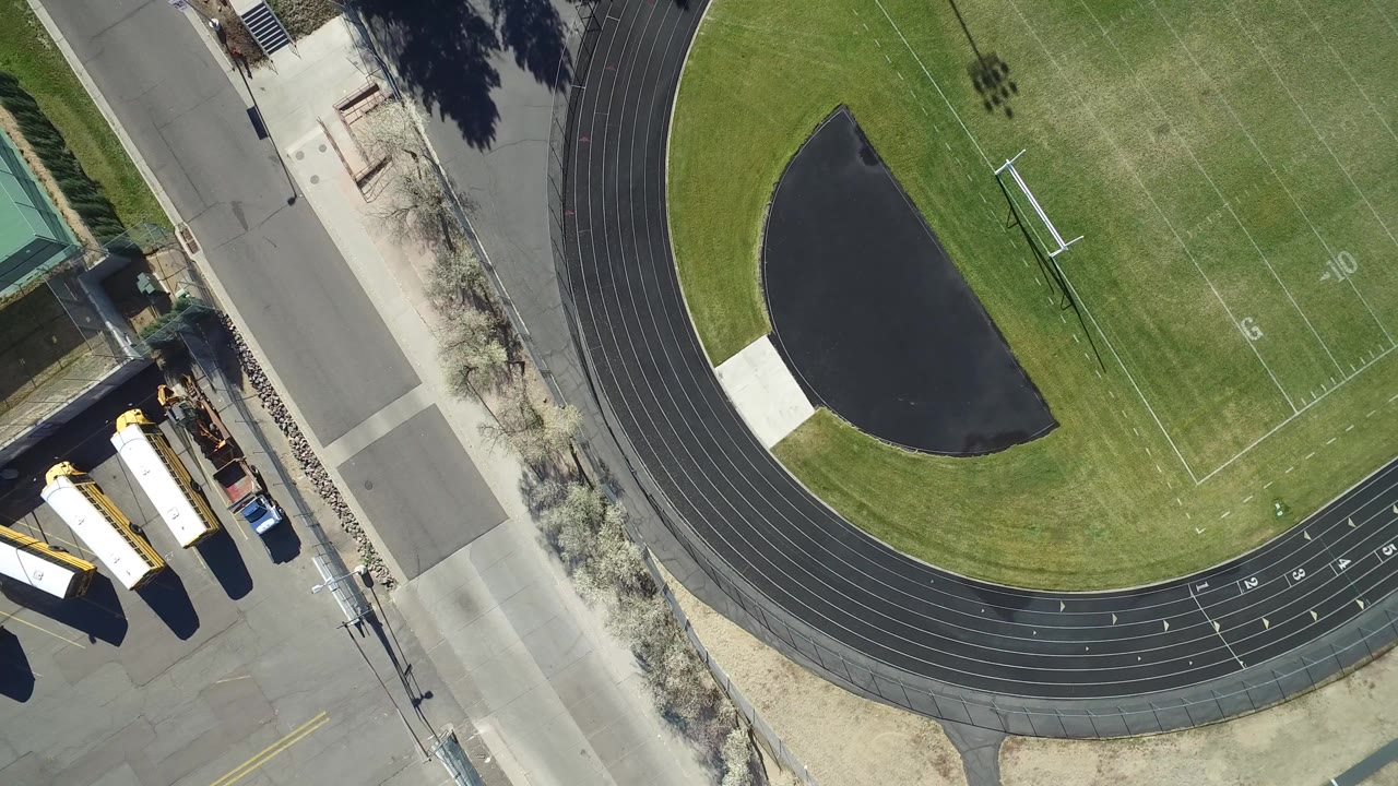 Straight-down aerial frame in Westminster, Colorado showing the curved black running track, part of the green football field, a road, sidewalks, parked yellow buses, and no visible people.