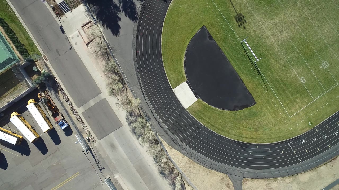 Overhead Westminster, Colorado view with the football field yard numbers, curved track lanes, dark paved infield area, adjacent road, and parked buses visible with no visible people.