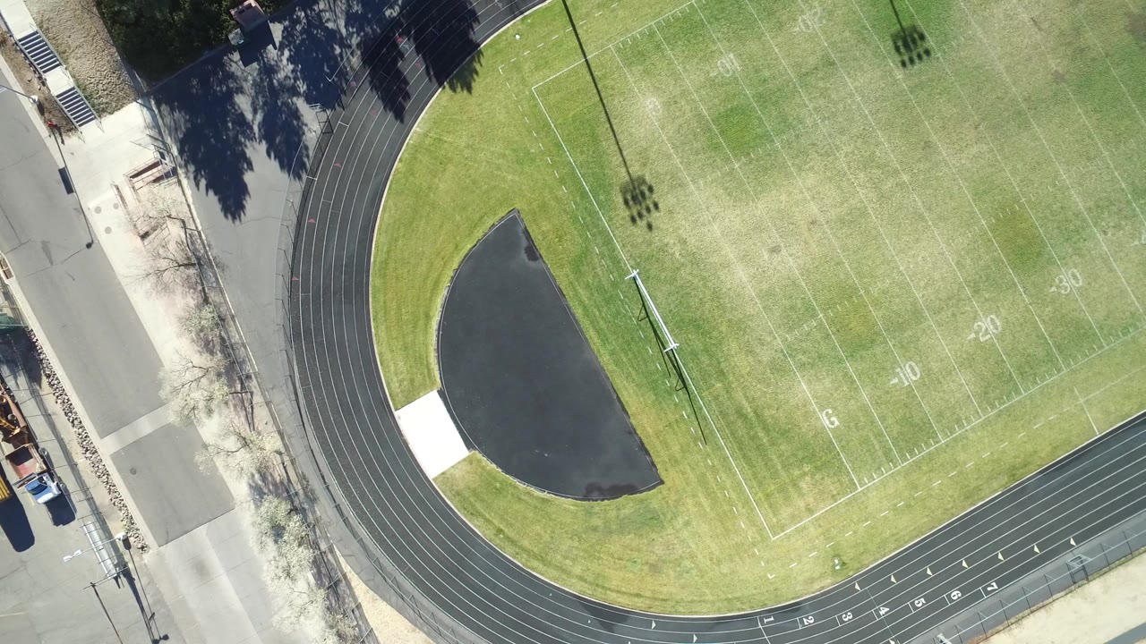 Overhead view in Westminster, Colorado of a lined green sports field, curved black running track, dark infield surface, road edge, and no visible people.