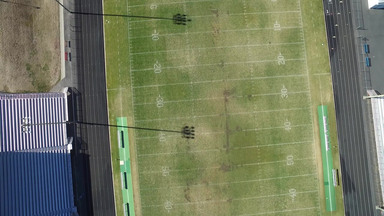 Straight-down aerial view of a Westminster, Colorado football field with yardage markings, a black running track, blue-and-white grandstands, light pole shadows, and no visible people.