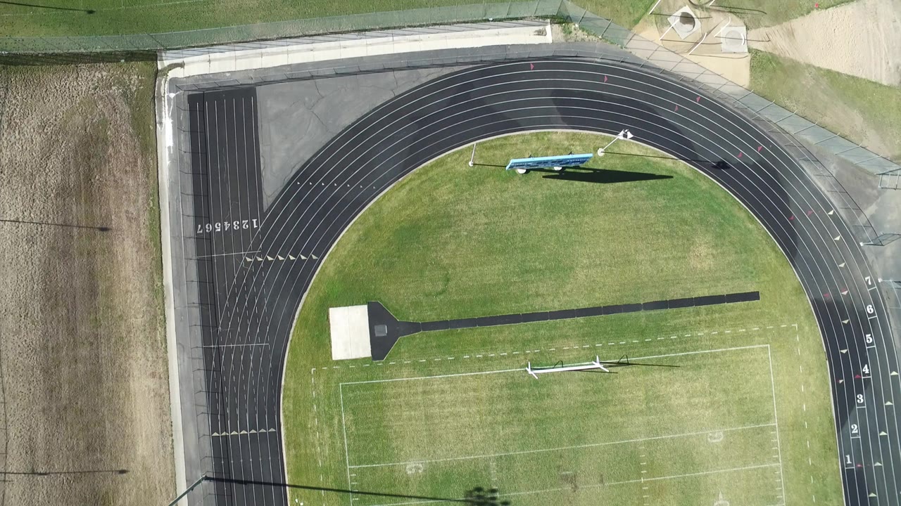 Overhead frame in Westminster, Colorado showing a black running track curving around a green football field with field equipment and no visible people.