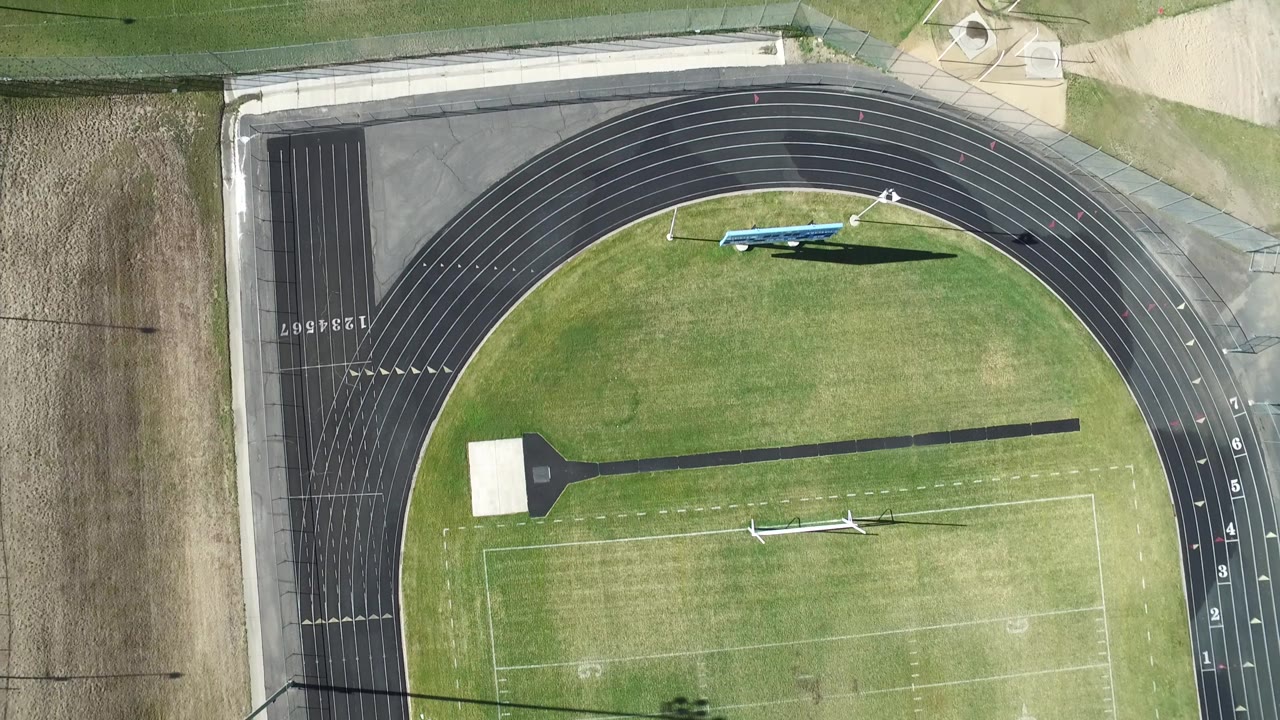 Aerial frame in Westminster, Colorado showing the curved track, grass field, painted markings, a long shadow from field equipment, and no visible people.