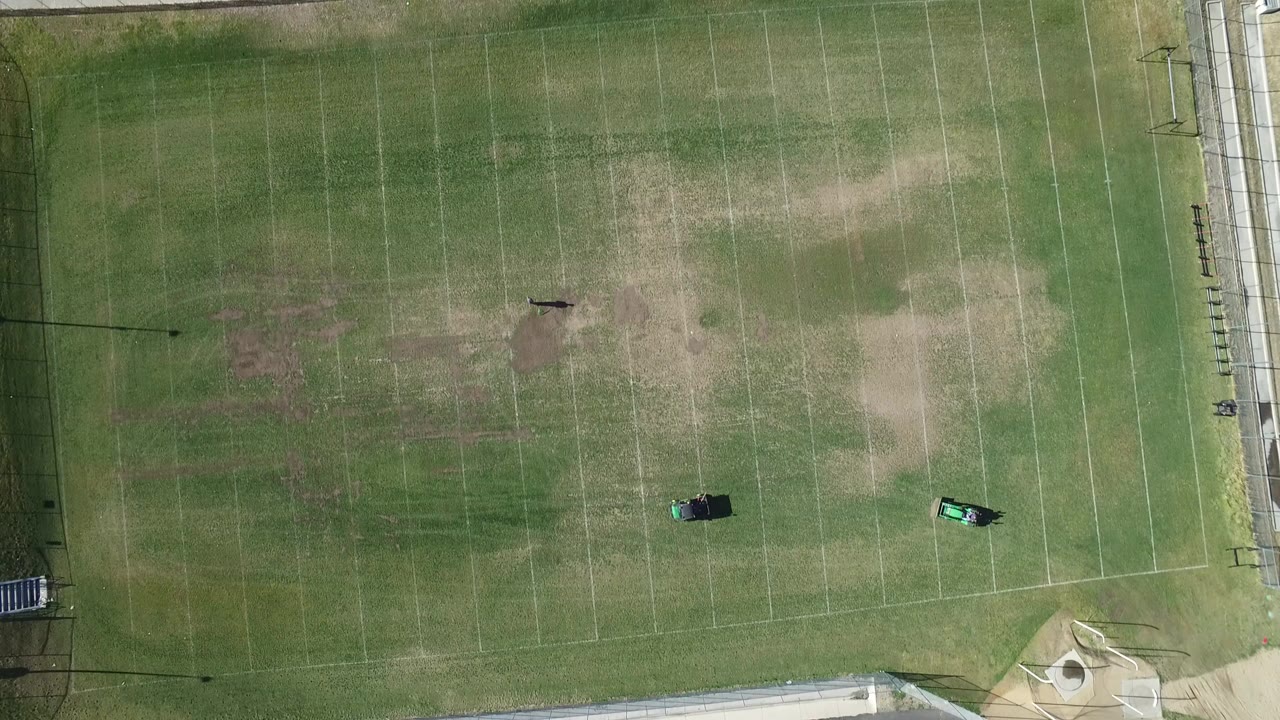 Overhead view of a marked green football field with worn patches and sideline fencing.