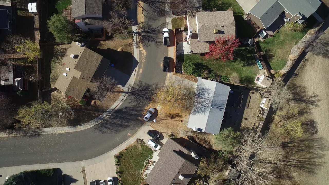 Aerial frame in Westminster, Colorado showing a curved neighborhood road, several house roofs, small lawns, parked vehicles, bare trees, and no visible people.