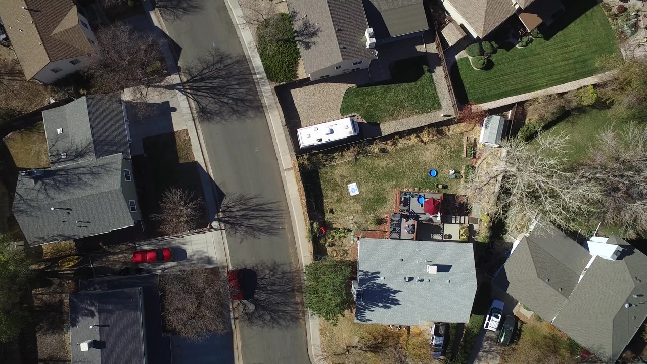 Top-down suburban block with rooftops, a street, yards, trees, and parked cars.