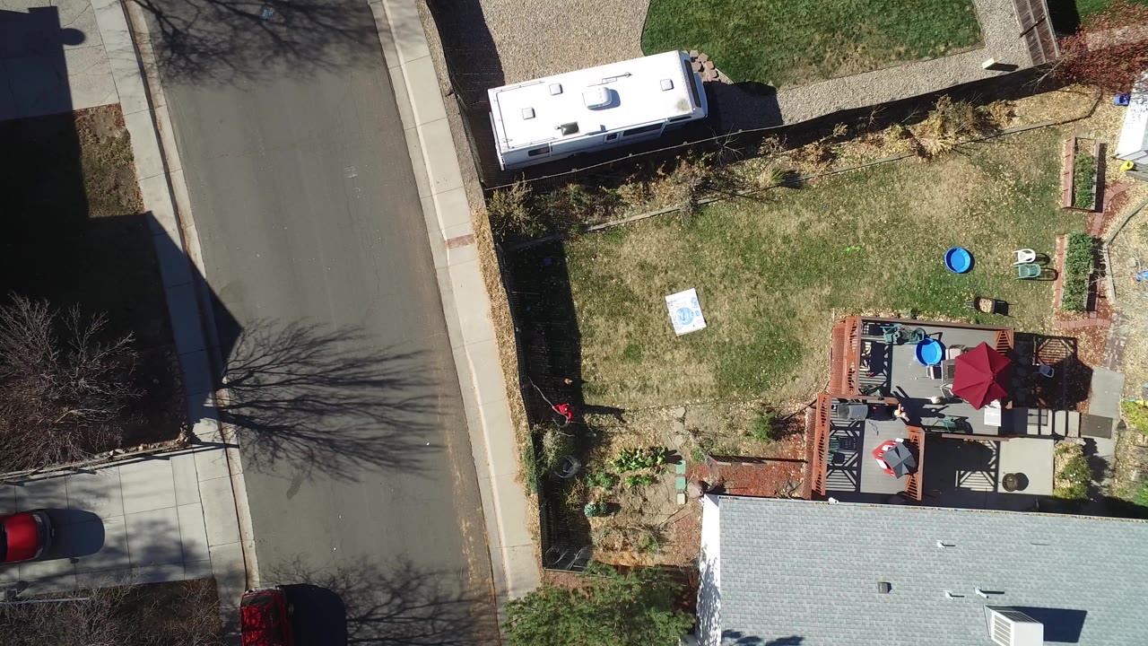 Overhead frame in Westminster, Colorado showing a residential road, sidewalk, lawn, gray roof, patio with red umbrella, white RV, and tiny people visible on the deck.